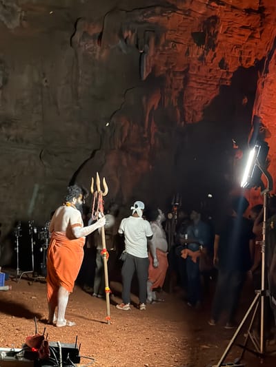 The image shows a dimly lit cave interior, possibly a film set, in Bethamcherla, India. A man, presumably an actor, stands facing away from the camera, dressed in an orange dhoti and adorned with white body paint and beaded necklaces. He holds a large, wooden trident with beaded embellishments. Several other people, appearing to be crew members, are visible in the background, some holding equipment, and others observing. Lighting equipment, including a bright spotlight, is set up to illuminate the scene. The cave walls exhibit rough, natural rock formations, and the ground is covered in reddish-brown dirt. The overall atmosphere suggests a production in progress within a natural cave environment.