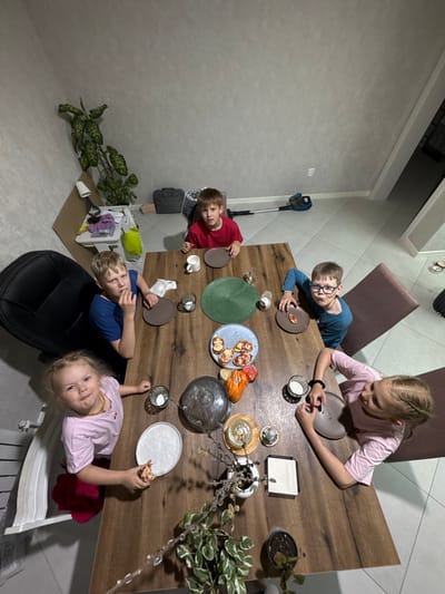 Certainly! Here's a detailed description of the image:
**Scene:** A top-down shot captures a group of five children gathered around a wooden table in what appears to be a dining area. The location is Ферма, Russia.
**People:** There are five children visible in the image:
* A girl with blonde braided hair, wearing a pink t-shirt
* A boy with glasses and a blue shirt.
* A boy with blonde hair and a red shirt.
* A blonde boy wearing a blue shirt.
* A young girl in a pink shirt with blonde hair holding a piece of pizza.
**Objects:**
* A large wooden dining table.
* An assortment of plates, some containing food (possibly small pizzas or appetizers).
* Various drinking glasses and cups.
* Several decorative objects: plants, candles, and other ornamental items.
* Chairs around the table, including a black chair and a purple chair.
* A vacuum cleaner standing against the wall.
* Various furniture and objects along the wall including plants, a small table, and a suitcase.
**Actions/Interactions:** The children seem to be in the middle of a meal or snack time. The young girl on the bottom is holding a piece of pizza. The other children are either eating, preparing to eat, or simply sitting at the table.
**Environment:** The setting appears to be a home dining area. The wall is painted in a neutral gray. There is a plant in the corner of the room.