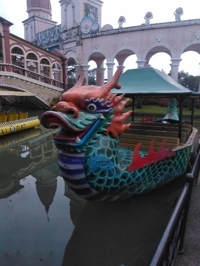 The image depicts an outdoor scene at the Parque de Diversiones, located in Ciudad de San José, Costa Rica. In the foreground and slightly right of center, a large, multi-colored dragon-headed boat floats in calm, dark water. The boat's head is sculpted with an orange-red base, featuring green, blue, and purple details, black eyes, white teeth, and prominent orange spikes/horns. Its body is patterned with teal-green scales, accented by red dorsal fins along the side, and contains light-colored wooden bench seats under a small, light green canopy.
To the left of the dragon boat, another yellow boat with "TAXI BOAT" stenciled in black on its side is visible, also floating in the water. The background is dominated by an elaborate architectural complex characterized by pale pink/peach walls, white columns, and multiple arches, reflecting a whimsical or themed design. A red-tiled conical roof of a tower is visible on the far left, alongside decorative light blue panels and circular motifs on the building facades. Several small, round ornamental figures are perched along the roofline of one section. A bridge with multiple arches extends over the water on the left, connecting different parts of the complex.
The still water perfectly reflects the background buildings and the overcast, light grey sky. A dark, ornate metal railing runs along the bottom right edge of the frame, separating the water from an unseen pathway. No individuals or specific actions are depicted within the scene.