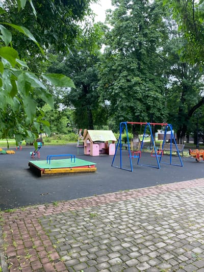 This is a photograph of a playground in the city of Zapadny, Russia. The playground is situated in a park-like setting with mature trees providing shade. The ground is covered in a dark, resilient surface, with a paved brick pathway bordering the area in the foreground.
The playground equipment includes a sandpit with a green lid and blue railings, a pink playhouse with a green roof, and a blue swing set with two red swings. In the background, there are several park benches and what appears to be a small amusement ride with cartoon-like characters. The foliage is lush and green, suggesting it is summertime or late spring. The overall scene depicts an empty, tranquil playground on an overcast day.