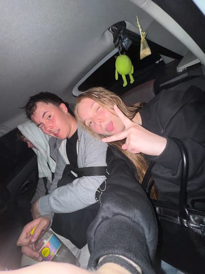 Three individuals are visible in the interior of a vehicle. The person in the foreground on the left appears to be a young male with dark hair, wearing a gray hoodie. He is looking towards the camera with one eye winking and his mouth open. To his right is a young female with long blonde hair, also facing the camera. She is sticking out her tongue and making a peace sign with her right hand. She is wearing a black hooded sweatshirt. Partially visible behind them on the left is another individual, appearing to be a young male, with a white towel draped over his head and shoulders. In the foreground, a plastic bottle with a colorful label that reads "A4" is being held by the person on the left. Hanging from the car's ceiling are two items: a bright green plush toy resembling a character with eyes and limbs, and a small golden pouch. The vehicle's interior appears to be dimly lit. There is no visible indication of the location being the city of Ryabinino, Russia.