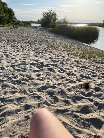 The image depicts a sandy beach adjacent to a body of water, likely a river or lake, under a bright, hazy sky. The foreground is dominated by the textured sand, marked with numerous footprints and some small clumps of dry vegetation. In the middle ground, several individuals are visible on the beach. One person lies on a towel, appearing to sunbathe. Further back, a small group of people are standing and walking. Beyond them, the water is bordered by a dense growth of tall reeds. Trees and foliage line the opposite bank of the water. The overall impression is a serene outdoor scene during daylight hours, suggesting a recreational area. The location is identified as Zhurivka, Ukraine.