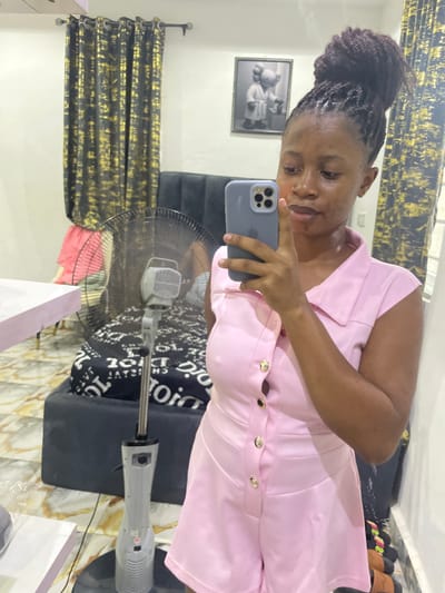 A female subject is depicted taking a mirror selfie indoors in Okpanam, Nigeria. The subject, a woman with brown skin and dark, textured hair styled in a high bun, is wearing a short-sleeved, collared pink romper with gold buttons down the front. She holds a light gray smartphone with multiple rear cameras to her face, capturing her reflection.
The background, visible in the mirror's reflection, includes a dark gray upholstered headboard or sofa. A black and white patterned sheet, possibly featuring the text "DIOR," covers a bed or mattress behind this furniture. A gray standing floor fan is positioned in the midground. On the wall to the right, a framed artwork displays two cartoon-like figures in a grayscale palette. Dark curtains with a gold abstract pattern are visible on the left side of the background. The floor features light-colored patterned tiles. A light pink surface, likely a table, is partially visible on the far left. Items resembling sandals are partially visible on the floor at the bottom right.