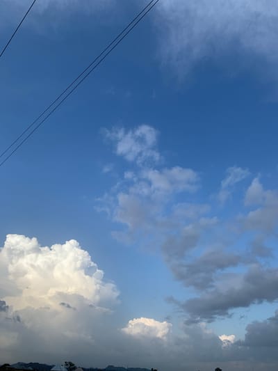 The image depicts a vast expanse of blue sky over Kuru Karama, Nigeria, with a dramatic display of clouds. Two parallel black wires stretch diagonally across the upper left quadrant of the frame, silhouetted against the bright sky. The clouds vary in form and color, ranging from bright white, puffy cumulus formations in the lower left to darker, more ominous gray clouds scattered throughout the mid-ground and right side. The lower edge of the image shows a dark, indistinct silhouette of a cityscape or rural landscape, with hints of buildings and trees, suggesting the geographical context of Kuru Karama. The lighting indicates it is either early morning or late afternoon, given the contrast between the illuminated white clouds and the shadowed areas. No people or specific actions are visible.