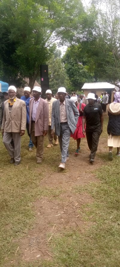 The image shows a group of ten men walking in a field in Burundu, Kenya. The men are dressed in suits and white caps, and they are walking in a single file line. The men are walking on a dirt path, and the field is covered in green grass. There is a large black speaker on the right side of the image, and a few people are visible in the background. The location is Burundu, Kenya, based on coordinates 0.459 latitude and 34.776 longitude. There are trees in the background.