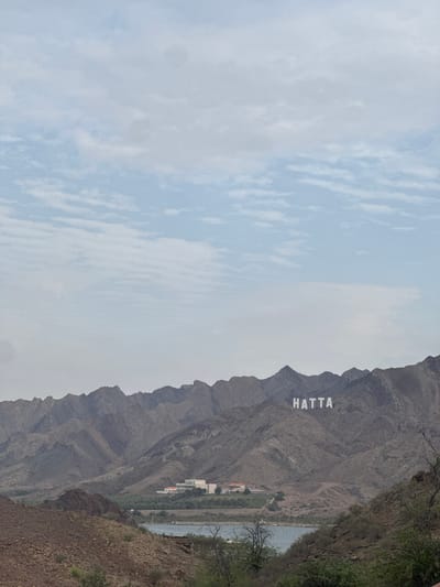 The image shows the "HATTA" sign placed on a mountain in Hatta, United Arab Emirates. The landscape includes a mountain range with the large, white "HATTA" sign prominently displayed on one of the peaks. Part of a body of water, potentially a lake or reservoir, can be seen along the side of the mountain. Buildings and greenery are visible on the mountain side, which is adjacent to the water. The sky has a mix of light blue and white clouds. The image has been rotated or cropped so that the landscape is on the right side of the frame and the sky fills the left side. The foreground on the right side is rocky and covered with low vegetation.