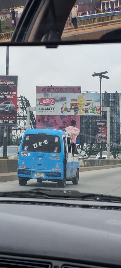 The image shows a street scene in Ikoyi, Nigeria. A blue and white mini-van with a person standing on its roof is driving on a road. The van has the word "OPE" written on its rear window. In the background, there are several billboards advertising various products, including "Laziz" quality food products and "Mikano International Limited". The sky is overcast.