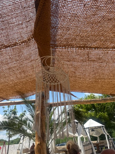 The image captures an outdoor seating area, likely a restaurant or cafe, in Kranevo, Bulgaria. The scene is dominated by a rustic overhead canopy made of brown, woven material, supported by wooden beams and a rough-hewn wooden post. Hanging from the post is a white, macrame-style decoration.

In the background, palm trees and lush green foliage are visible against a bright blue sky with scattered clouds. Several white, tent-like structures, possibly cabanas or gazebos, are set up, providing shade and enclosure for seating areas. Tables and chairs are visible within these structures.

A person is seated at a table within one of the cabanas, appearing to be relaxed. In the foreground, the back of a person's head with blonde hair is visible, suggesting they are also a patron of the establishment. The overall atmosphere appears to be casual and summery, with an emphasis on natural materials and open-air dining.