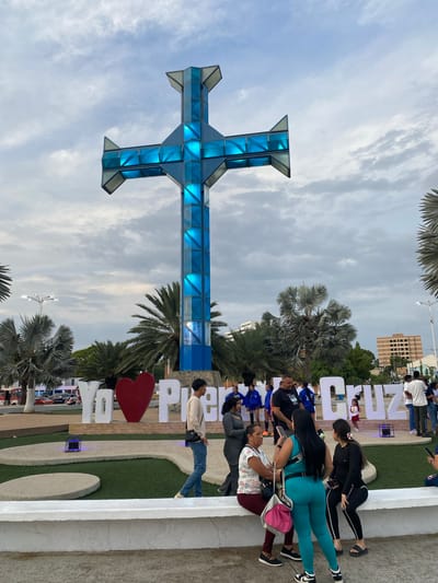The image shows a public space in Puerto La Cruz, Venezuela. A large, blue, geometrically designed cross stands prominently. People, including adults and children, are interacting and posing on a low, white structure with the city name spelled out in large letters behind it. There are palm trees and other greenery around the cross and lettering. The sky is cloudy. A building can be seen in the background behind the crowd and letters.