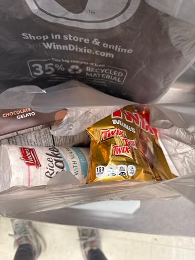 A bag of groceries contains a bag of Twix Mini chocolate candy, a package of Wieber's thin rice cakes with sea salt, and chocolate gelato. The plastic grocery bag has "Shop in store & online SnoWinnDixie.com" printed on it, indicating the store where the items were purchased. The bag also specifies it contains at least 35% recycled material. The nutritional information on the Twix bag shows 150 calories per 3 pieces.