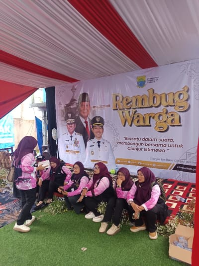 This image captures an outdoor event under a tent structure in Kawungluwuk, Indonesia. The tent's ceiling is adorned with draped red and white fabric panels. The ground is covered with artificial green turf in the foreground, transitioning to a patterned mat further back. A large white banner acts as the backdrop, displaying "Pemerintah Kabupaten Cianjur" (Government of Cianjur Regency) with its logo, the words "Rembug Warga" (Citizen Deliberation) in golden script, and the slogan "Bersatu dalam suara, membangun bersama untuk Cianjur Istimewa." The banner features images of four men: a prominent central figure, and three men in uniform below him, identified by names such as "H. Herman Suherman," "TB. Mulyana Syahrudin," and "Dr. H. Wawan." The banner also includes social media icons and "cianjurkab.go.id."
Eight women, all wearing hijabs, are present. Most are dressed in light-colored, long-sleeved shirts under dark vests and dark pants, many with ID badges. One woman stands on the left, holding a white object in her hands, facing a group of seven women who are seated or squatting on the ground. The seated women appear to be observing, with one individual looking at a mobile phone. White flowers are arranged along the base of the banner. A cardboard box containing multiple small plastic cups of liquid is visible on the far right. A wooden object is partially visible on the far left. An exterior building and a blue tarp are discernible in the background.