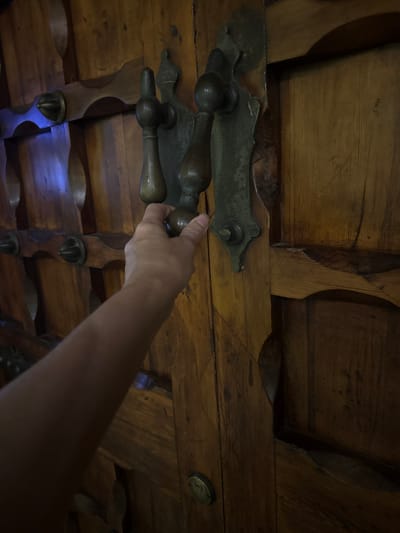 The image shows a close-up, low-angle view of an antique wooden chest, likely in Sevilla, Spain. The primary subject is a person's hand reaching out to touch one of the chest's ornate, brass handles. The chest itself is constructed from dark, aged wood with visible grain and some wear. It features several decorative elements, including carved wooden supports and large, metallic hardware that serves as both handles and decorative accents. The hand, belonging to an unseen person, is positioned to grasp a rounded, bulbous handle. The scene is dimly lit, suggesting an indoor setting, possibly a dimly lit room or a cellar. The overall impression is one of historical richness and tactile engagement with an antique object. No text is visible in the image.
