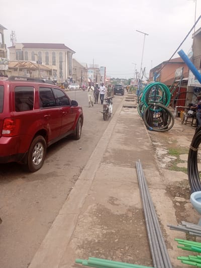 In the city of Dadi Riba, Nigeria, a roadside scene unfolds under an overcast sky. A red SUV is parked on the left side of a paved road, its passenger side facing the viewer. A concrete sidewalk runs parallel to the road, separating it from a collection of materials. On the sidewalk, bundles of green and gray pipes are stacked, alongside coiled green hoses and what appears to be a light blue toilet bowl. Further down the sidewalk, a person is partially visible in the background, seemingly working at a stall or shop.
Across the road, several individuals are walking, with one person on a motorcycle moving away from the camera. In the distance, buildings line the street, including a large, light-colored structure with arched windows and a prominent antenna tower. Traffic, including cars and a tricycle, is visible on the road, suggesting active movement within the urban environment. The overall impression is one of a bustling commercial street with construction or plumbing supplies being displayed for sale.