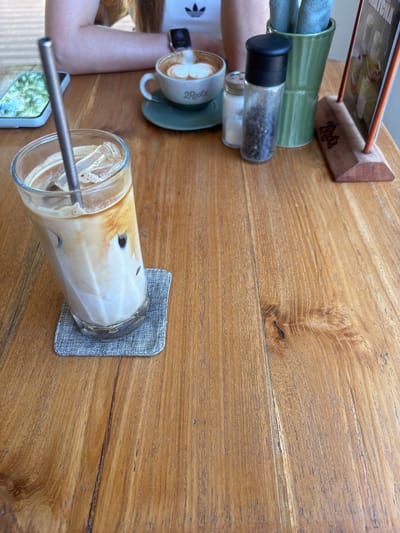 The scene is captured on a wooden table, likely in Pecatu, Indonesia, given the presence of "2 Roots" branding, which is associated with a café in that area. The table features drinks: a glass of iced coffee with a metal straw, and a cup of latte with milk art on a green saucer. The coffee is accompanied by condiments such as salt and pepper shakers, and a napkin holder. There is also a phone. An arm is visible, clad in a white Adidas t-shirt. The composition is shot from an overhead angle.
