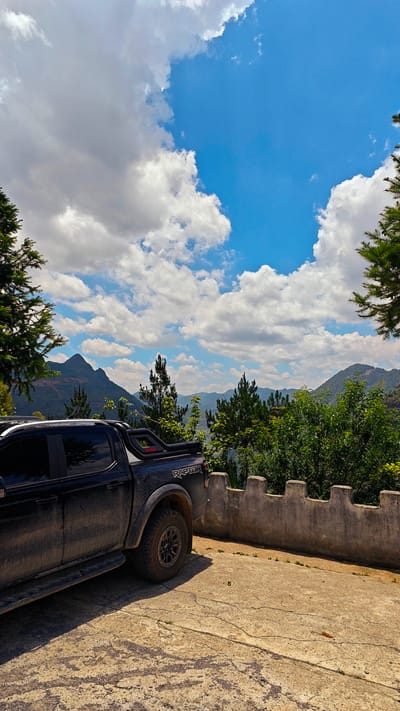 The image shows a black Ford Ranger Raptor truck parked on a concrete surface in the Lũng Cú Commune, Vietnam. The truck is partially obscured by its own dirt and the left side of the frame, but its "RAPTOR" badging is visible. A low, crenellated concrete wall runs along the edge of the parking area, beyond which are trees, shrubs, and a mountainous landscape under a sky filled with clouds. The environment is indicative of a rural, elevated location within the commune. There are no visible people. The scene suggests an outdoor setting, possibly a viewpoint or overlook.