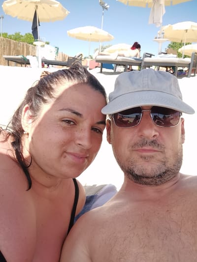 This is a selfie taken by a couple on a sunny day at the beach in Kranevo, Bulgaria. The man, wearing a light grey baseball cap and sunglasses, is positioned in the foreground on the right. He has a short, grey-flecked beard and is looking towards the camera. The woman, on the left, is also looking at the camera. She has dark hair pulled back and is wearing a black bikini top. In the background, there are beach chairs, umbrellas, and other beachgoers lounging in the sun. The sand is visible in the foreground and stretches towards the sea. The sky is clear and blue.