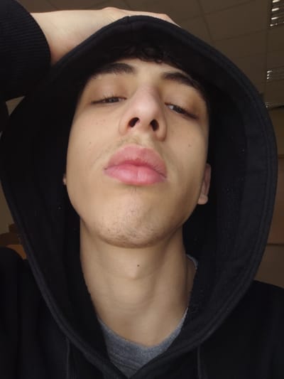The image depicts a close-up portrait of a young male subject located indoors in Jibb as-Safa, Syria. The subject is a young male with light to medium skin tone, dark curly hair visible beneath a raised black hooded sweatshirt, and sparse dark stubble present on the upper lip, chin, and jawline. His dark eyes are partially lidded and appear to be looking directly at the camera. His lips are full and slightly parted, appearing a pinkish hue. The right earlobe (viewer's left) shows a small, dark stud or earring. The subject's left hand (viewer's right) is raised, with fingers visible resting on the top of the black hood. He wears a black hooded sweatshirt over a grey crew-neck t-shirt. The background is an indistinct, blurry indoor environment featuring light-colored ceiling tiles with visible seams and what appear to be light fixtures at the top, and light-colored walls on the viewer's left. The lighting is artificial, originating from above.