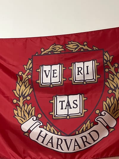 The image shows a close-up of a Harvard University flag. The flag is red and features the university's crest in the center. The crest is enclosed by a golden laurel wreath. Within the crest are three stacked books, each with Latin text on them. The left book has "VE" and "RI" stacked vertically. The middle book has "TAS" stacked vertically. A banner curves around the right side of the crest, bearing the text "HARVARD" in large, black, serif letters. The flag appears to be made of fabric and is slightly creased. The background is a plain, light-colored wall, suggesting the flag is displayed indoors or against a neutral backdrop. The image is taken at a slight angle, with the right side of the flag closer to the viewer than the left. There are no people visible in the image, and the focus is solely on the flag and its details. The setting is not clearly identifiable beyond the flag itself and the plain background, although the prompt provides a location context of Daxing District, China, this context cannot be inferred from the visual information alone. The image appears to be a still photograph, with no indication of a specific time of day or weather conditions. The emotion conveyed is one of institutional pride or affiliation, represented by the university's emblem.