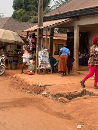 The image depicts an outdoor commercial scene in Otuocha, Nigeria. The foreground features an unpaved reddish-brown dirt road, uneven with depressions and a shallow drainage channel containing debris on the right. In the midground, a wooden utility pole stands centrally with power lines extending across the frame.
On the left, a market stall operates under a light-colored umbrella canopy, displaying fruits and vegetables in baskets on a wooden stand. A silver motorcycle's front wheel is visible on the far left. Adjacent to the market stall, a woman with dark hair, wearing a white top patterned with red and blue (resembling the US flag) and white shorts, stands barefoot, her left arm raised. A large woven sack and an orange crate rest near her feet. Behind her, a rack displays various clothing items.
Centered in the midground is a blue and white "CYNTHIA POS" sign, advertising financial services such as withdrawals, transfers, deposits, and data/airtime sales, with a phone number (09136198207) displayed. The sign is propped on a wooden easel.
To the right of the sign, a light-colored building with an open doorway reveals two individuals seated inside. Outside this building, a woman wearing a light blue top and a long reddish-brown skirt walks away from the camera, towards the building's entrance. Several plastic chairs, including green and purple ones, are positioned near the building's entrance. Further right, a woman with bright red curly hair, wearing a plaid shirt and bright pink leggings with black slides, walks from right to left, partially obscured.
The background shows multiple structures with corrugated metal roofs, some rusted brown and others grey, along with tropical foliage and palm trees under an overcast sky.