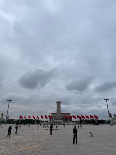 The image depicts Tiananmen Square in Dongcheng District, China. A tall stone obelisk stands near the center, flanked by rows of red flags on either side. Several people are present in the square, with some walking and others standing. The ground is paved with large stone slabs. Tall light poles are positioned along the edges of the square. The sky is overcast with heavy clouds.