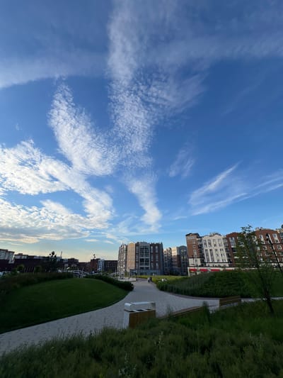 The image depicts a contemporary urban landscape with modern apartment buildings and a park area in the city of Pervomaysky, Russia. The sky is a vibrant blue, populated by scattered white clouds forming wispy patterns. In the foreground, a curved pathway made of light-colored paving stones winds through a well-maintained park. Lush green grass covers a gently sloping mound to the left of the path, and dense, tall green grass borders the lower edge of the frame.

Several multi-story apartment buildings with varied facades, including brick and lighter-colored materials, dominate the background. They feature multiple windows and balconies. Some of the buildings have red awnings. A few people can be seen walking or standing in the distance within the park area. The park also contains benches and planters. The overall scene suggests a residential area with public green spaces designed for leisure and community. The lighting indicates it is daytime, possibly late afternoon or early evening due to the soft light.