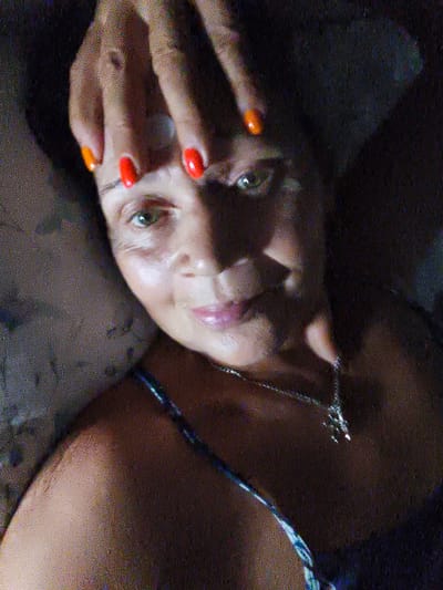 This is a close-up, grainy, indoor portrait of a woman. Her face is partially obscured by her hands, which are resting on her forehead. Her fingernails are painted in bright red and orange hues. The woman has green eyes and is looking towards the camera with a slight smile. She is wearing a dark top with a blue and white pattern and a silver necklace with a cross pendant. The lighting is dim, suggesting it might be nighttime or a dimly lit room. The background is dark and indistinct, offering no specific setting cues. There is no visible text in the image. The overall mood appears relaxed and perhaps playful, given the brightly colored nails and the woman's expression.
