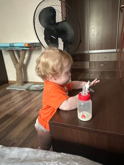 A toddler with blond hair is standing next to a dark wooden table, reaching for a baby bottle with a red cap. The toddler is wearing an orange t-shirt and a diaper with white lace trim. A pacifier is in the toddler's mouth. In the background, a black fan is visible, along with a cat tree and wooden cabinets. The scene appears to be indoors, in a residential setting in Sayanogorsk, Russia.