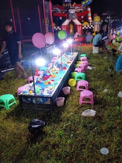 A nighttime scene in Kecamatan Sambirejo, Indonesia (latitude: -7.49, longitude: 111.119), featuring a fish-catching game. The game is set up in a grassy area illuminated by lights. A long, rectangular tank filled with water and artificial fish is surrounded by plastic stools of various colors (pink, green, and yellow) placed in rows.  A man is standing nearby. The ground is covered in green grass. Two pink balloons are floating in the air. A large inflatable structure resembling a play area is visible in the background. There are also several baskets and plastic cups scattered around the area.  A black plastic bag is laying in the grass.  A white plastic cup is laying on the ground.