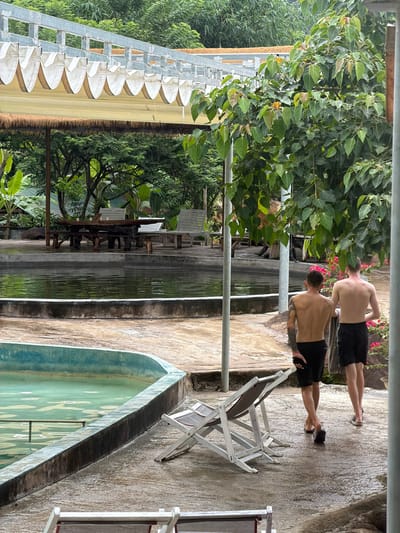 Two shirtless young men, one with a tattoo on his left arm, walk away from the camera on a wet concrete path. The man on the left wears black shorts, and the man on the right wears black shorts with white stripes. They are in an outdoor setting with lush green foliage surrounding a pool and a pond. In the background, a wooden picnic table with benches is visible next to a body of water. Two white lounge chairs are on the concrete path. The environment appears to be a resort or natural park in Khanh Vinh, Vietnam, with the architecture and vegetation suggesting a tropical or subtropical climate. The lighting indicates it may be overcast or late afternoon.
