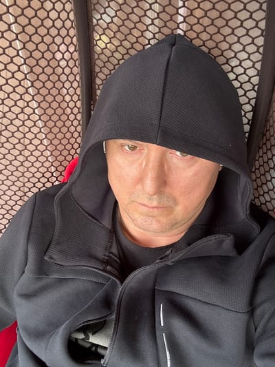 A man wearing a black hooded jacket is shown in a close-up, selfie-style photograph. The background consists of a textured, honeycomb-patterned mesh in a reddish-brown color. The man's face is partially obscured by the hood of his jacket. He has light skin, brown eyes, and is looking directly at the camera with a neutral expression. The image does not provide any information about a location in Gavshino, Russia.