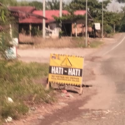 This blurry daytime image captures a rural or semi-rural roadside scene in Tanjongkeumala, Indonesia. The central focus is a yellow temporary warning sign positioned on the dirt shoulder beside a paved road, which curves to the right in the background.
The warning sign features a black border and bold black text on a yellow background. The prominent text reads "HATI - HATI," which translates to "BE CAREFUL" or "CAUTION." Above this text, a black warning triangle symbol is visible. Below the main text, smaller black lettering appears to state "50 METER KE DEPAN ADA PEKERJAAN JALAN," indicating "50 METERS AHEAD THERE IS ROAD WORK." The sign rests on a wooden frame.
To the left and foreground of the sign, green grass and foliage are present. The road surface appears somewhat unkempt, with patches of dirt and possibly some debris. In the blurry background, several single-story buildings with reddish-brown roofs are visible amidst trees and other vegetation, suggesting a residential or village setting. Utility poles are also scattered throughout the background. The overall impression is a notice for drivers about upcoming road conditions in a moderately developed area.