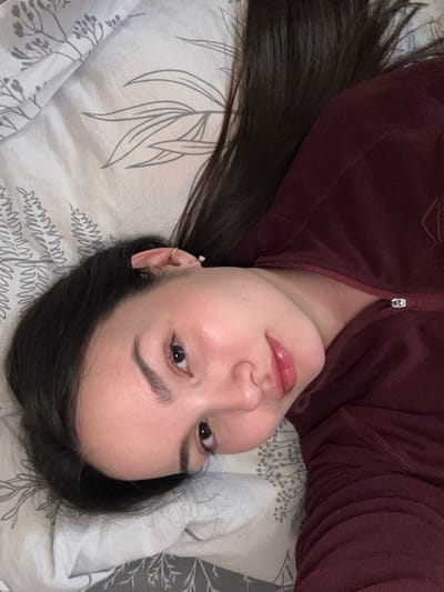 A single female individual is depicted in an indoor setting within Bayronovka, Russia. The subject is positioned diagonally, lying on her back and looking directly into the camera. She has long, dark brown hair spread across a light-colored surface with a grey botanical pattern, likely a bedsheet or pillow. Her face features fair skin, dark eyebrows, and dark eyes. Her lips are slightly parted and appear glossy with a light pink tint. The visible left ear has two helix piercings: one with a small gold-colored hoop and another with a small stud. She is wearing a dark reddish-brown garment, possibly a zippered fleece or hooded sweatshirt, with a portion of a silver-toned zipper visible on the right side of the frame.