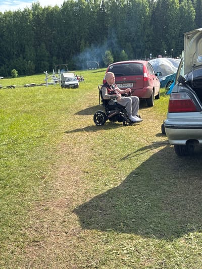 The scene depicts an outdoor gathering in a grassy field in Большая Кивара, Russia, under clear daylight conditions. In the mid-foreground, a light-skinned male with a shaved head and tattoos on his left arm sits in an electric wheelchair. He is wearing a dark t-shirt with red patterns, light grey sweatpants, and white socks or shoes, holding a control device for the wheelchair. He is positioned facing towards the left.
Behind the man, a red Lada Kalina hatchback is parked, displaying Russian license plate `C 414MO 18`. To the right of the man, the rear of a silver sedan with an open trunk is partially visible. Further into the midground, to the left of the red car, a white pickup truck is parked near a metallic structure of white poles. Behind these vehicles, a large white tarp-covered object is present. Smoke is observed rising from the left side of the midground, indicating a burning source.
The ground is covered in green and dry grass, with a well-trodden dirt path running diagonally from the bottom left corner towards the center. Long shadows are cast from the right side of the image towards the left. In the background, a dense line of tall green deciduous trees forms the horizon. Partially visible within the trees are two wooden swing sets, one towards the left and another towards the right. The sky above is light blue with sparse white clouds.