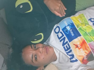 The image shows a woman lying down next to a large black stuffed toy with yellow eyes, possibly a dragon or monster. She is wearing a white t-shirt with a colorful graphic print on it. The t-shirt has text in Spanish that reads "Hoy te veo" and "Fan de mi". The woman has dark curly hair and is looking towards the camera with a neutral expression. Her right hand is resting on the stuffed toy. The background is blurred but appears to be indoors, possibly a bed or couch with a green blanket. There is no clear indication of the location in El Valle del Espíritu Santo, Venezuela.
