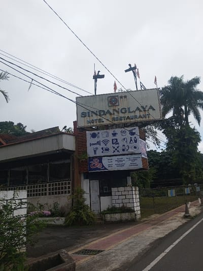 An outdoor scene captures the front facade of the Sindanglaya Hotel Restaurant building in Thotagamuwa, Sri Lanka, under an overcast sky. A prominent white sign spanning the upper portion of the structure displays "SINDANGLAYA HOTEL RESTAURANT" in dark blue capital letters. Above this sign, two flagpoles with orange flags are visible. Below the main sign, a multi-panel white sign features various service icons, including a bed, Wi-Fi symbol, parking 'P', laundry, food (bread, coffee, fries, noodles), fruit, and a fried egg, alongside text that includes "Malio S3 Cafe". The building itself presents a multi-tiered roofline, sections with horizontal white siding, and large windows. The lower right section of the building's front features a white brick facade. Numerous utility lines crisscross the sky above the building. To the left and behind the building, dense green foliage is visible, with a tall palm tree prominent on the right side. In the foreground, a gray paved sidewalk with a reddish-brown edge runs alongside a dark asphalt road. A black metal fence, partially draped with blue and white striped fabric, extends along the property line to the right. No individuals are present in the frame.