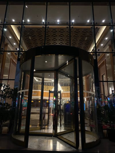 The media file captures a nighttime view of the brightly lit interior of a modern building, likely a hotel or commercial establishment in Doha, Qatar, seen through a large glass facade and a revolving door. The perspective is from outside, looking into the warmly illuminated space.
The central subject is a polished revolving door, constructed from glass panels and a metallic frame with a brass or gold-toned finish. This door is set within an expansive glass wall, composed of large rectangular panes connected by dark frames and circular spider fittings. Outside, the environment is dark, with faint outlines of potted plants visible on either side of the entrance.
Inside, the building's lobby or reception area is well-appointed. Numerous recessed spotlights illuminate a multi-layered ceiling with ornate cornices and reflective surfaces. A distinctive design element is a tall, dark wooden wall featuring a repeating geometric pattern of interlocking 'X' shapes. Further inside, a shimmering chandelier reflects off the glass, adding to the opulent ambiance.
In the lower right section, visible through the revolving door, is a reception desk with the word "RECEPTION" clearly displayed on a white sign. Near this area, a vertical banner or screen reflects the text "QATAR" and potentially parts of a "NIKE" logo, suggesting promotional material or branding within the building. The interior floor appears to be polished marble or a similar high-end material. No people are distinctly visible, maintaining a serene and unoccupied atmosphere. The scene exudes sophistication and grandeur, characteristic of contemporary architecture in the region.