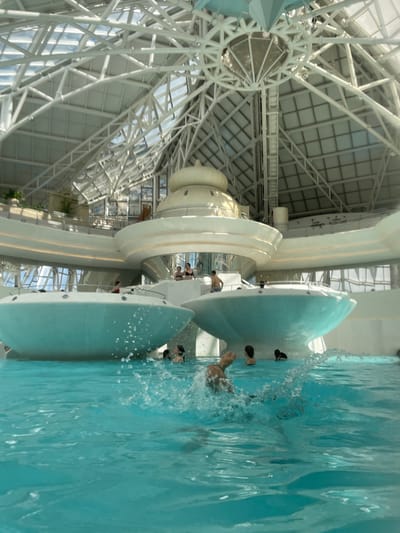 This indoor shot depicts the Caldea thermal spa in Les Escaldes, Andorra. The image captures a large, turquoise pool where people are bathing and swimming. There appear to be waterslides with water splashing in the pool. The architecture features a soaring, white metal framework that forms the ceiling, with glass panels allowing natural light to enter. White structures that appear to be part of the spa's design are also visible.