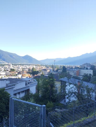 The image depicts a panoramic view of Muralto, Switzerland, on a clear, bright day. The foreground is dominated by a grey metal fence, likely part of a property boundary. Beyond the fence, the roofs of several modern buildings are visible, some with white facades and flat, grey roofs. Green trees and lush vegetation are scattered throughout, indicating a well-maintained urban environment with ample greenery.
In the midground, a town with a mix of residential and commercial buildings stretches out. The architecture features a variety of styles and colors, with some buildings having light-colored facades and others in warm earthy tones. A significant body of water, likely Lake Maggiore, is visible in the distance, with the city extending along its shores.
The background is defined by a majestic mountain range. The mountains are covered in dense green forests and rise sharply against the clear blue sky. The sunlight casts a soft glow on the mountains, highlighting their contours and creating a serene atmosphere. A thin contrail from an airplane is visible in the upper left portion of the sky, suggesting air traffic. The overall impression is one of a picturesque and peaceful lakeside town nestled against a dramatic mountain backdrop.