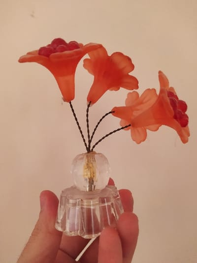 A human hand is shown in a close-up, holding a small, artificial floral ornament against a plain, light-colored background. The ornament consists of three translucent, bell-shaped flower components, reddish-orange in hue, each containing a cluster of small, red, spherical beads. These flower components are attached by thin, dark-colored wire stems to a clear, faceted glass base. The base has an upper spherical section with a visible yellow internal element, connected to a lower, wider, fluted glass stand. The hand grasps the lower part of this glass base. The specific location in Harasta, Syria, is not visually discernible within the image.