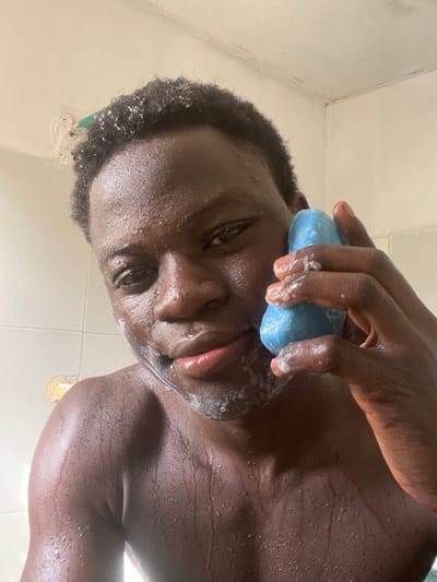 A dark-skinned individual, appearing male, is depicted in a close-up, chest-up shot within what appears to be a bathroom in Mosafejaw, Nigeria. The person has short, dark, curly hair, visibly wet and adorned with water droplets, similar to the skin across the face, neck, and upper torso. White soap suds are present on the chin and around the mouth area. The individual holds an irregularly shaped, solid blue bar of soap in their right hand, positioned against the right side of their face, near the cheek/jawline. Soap suds are also visible on the blue bar and the fingers of the right hand. The background consists of white ceramic tiles, with visible grout lines and some discoloration or dirt on the upper walls and ceiling edge, including what may be peeling paint or grime, and a small segment of a green object or pipe on the upper left. A small yellow object is partially visible on the lower left background tile. The person is looking directly forward, towards the camera.