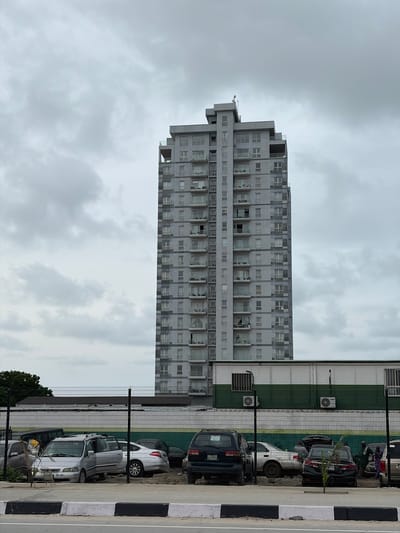 The image captures a static, vertical composition dominated by a tall, grey, multi-story building on the left side, set against an overcast, cloudy sky. The building appears to be an apartment block or a commercial structure with numerous windows and balconies. Some windows on various floors show signs of damage, with broken glass or missing panes, and some balconies appear cluttered or in disrepair.
To the right of the building, separated by a wire mesh fence, is a parking area. Several cars are parked here, including dark-colored SUVs and lighter-colored sedans. One light-colored car in the middle appears to be an older model, while a dark SUV in the background has a partially visible license plate reading "EPE 811...". Behind the fence and cars, there is a low-rise structure, possibly a garage or utility building, painted in horizontal stripes of dark green and white, with air conditioning units mounted on its exterior.
In the immediate foreground, a black and white striped curb or barrier is visible, suggesting a road or pedestrian path. A few sparse trees or bushes can be seen near the fence line at the bottom. The sky is uniformly grey and cloudy, indicating an overcast day. The overall scene suggests an urban or suburban environment in Ilado, Nigeria. There is no visible human activity, and the scene is characterized by the architectural presence of the building and parked vehicles.
