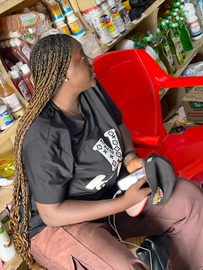 The image shows an adult female seated in an indoor commercial space, likely a shop or storage area, in Samaru, Nigeria. The subject has dark skin, long black and golden-brown micro braids, and is depicted in a profile view looking towards the right. She wears a black short-sleeved t-shirt featuring a large white 'V' logo with floral patterns, brown cargo pants, and a black shoe with a silver chain accent. A silver stud earring is visible on her right earlobe, and she wears a thin silver necklace and a dark bracelet on her left wrist. In her left hand, she holds a white smartphone connected by a white cable and a black baseball cap with a red brim and a yellow/red logo. She is seated on a red plastic chair, partially visible. The background features wooden or cardboard shelves densely stacked with various consumer goods, including multiple bottles of liquids (e.g., 'Hypo,' 'Viron,' green bottles of dish soap or similar products), a container of cotton buds, and a cardboard box labeled 'Sure Multipurpose Wipes.'