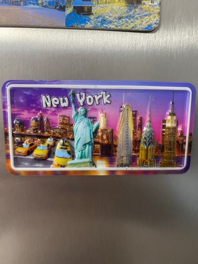 The image shows a rectangular magnet with a three-dimensional depiction of the New York City skyline. Prominently featured is the Statue of Liberty, rendered in a light blue-green hue, with its torch raised. Behind her, a vibrant cityscape unfolds, showcasing iconic skyscrapers like the Empire State Building and the Chrysler Building. The skyline is set against a dramatic purple and orange gradient sky, suggesting either a sunset or sunrise. Several yellow taxis are depicted on a street level below the Statue of Liberty. The text "New York" is visible on the magnet in large, raised white letters. The magnet is attached to a metallic surface, likely a refrigerator. To the left of the New York magnet, a portion of another magnet is visible, featuring a colorful, abstract design with blue and yellow elements, possibly depicting a landscape or artwork. The overall scene is static, with no discernible activity or event taking place beyond the representation of a New York scene. The location context provided is Łódź, Poland, however, the content of the magnet itself clearly depicts New York City.