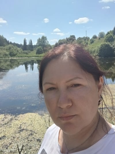 In a close-up selfie, a woman with reddish-brown hair and light skin faces the camera with a neutral expression. She is wearing a white t-shirt, a silver necklace with a small pendant, and hoop earrings. Her right eye is visible, and her hair is slightly windblown. Behind her, a body of water, likely a lake or pond, is visible, with reflections of trees and the sky. The water's edge is lined with vegetation, some of which appears to be aquatic plants. In the background, rolling green hills covered in trees and bushes rise under a clear blue sky with scattered white clouds. The overall scene suggests an outdoor, natural environment, possibly in a rural area.
