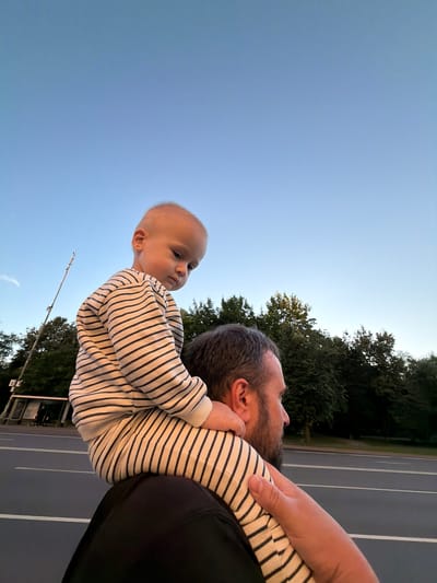 A medium shot captures a father and child from behind and side, with the child sitting on the father's shoulders. The father, with a beard and dark hair, wears a black shirt. The child, facing away from the camera and looking down, wears a striped long-sleeved shirt and pants. They are outdoors, with a clear blue sky above. In the background, a line of green trees runs horizontally. A road with white lines is visible in the mid-ground, and a bus stop with people can be seen on the left side of the image. A tall pole with antennas is also visible to the left of the trees. The scene appears to be in Moskovskiy, Russia.