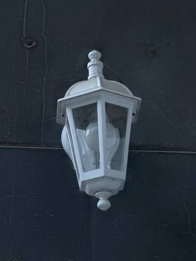 This is a close-up, overhead shot of a white outdoor lantern. The lantern has a rectangular frame with clear glass panes and a visible light bulb inside. It is mounted on a dark, textured surface that appears to be stone or tile, possibly part of an exterior wall or pavement. The surface has visible seams and circular indentations, suggesting it's made of multiple panels or sections. The lighting suggests it is daytime, but the overall tone is muted, possibly indicating overcast conditions or shade. The image has a dark, cool color palette. There is no visible text. The setting is suggestive of an outdoor area in Athens, Greece, perhaps a courtyard, walkway, or building exterior. The focus is solely on the lantern and its immediate surroundings, with no people or other discernible objects.