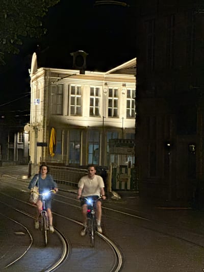 This image depicts a nighttime scene in Ghent, Belgium, specifically on a street featuring tram tracks. Two individuals are riding bicycles along the tram tracks. The person in the foreground on the left is wearing a blue denim jacket and a light-colored bag is slung over their shoulder. The person on the right is wearing a white t-shirt and dark shorts. Both bicycles have illuminated headlights.
The background features a brightly lit building with multiple windows, suggesting it is a commercial establishment or residence. A yellow awning or cloth is visible hanging from the building. The street is paved and the tram tracks are prominent. The overall atmosphere is dark, typical of a night scene in an urban environment. The image is slightly blurred due to motion and low light conditions.