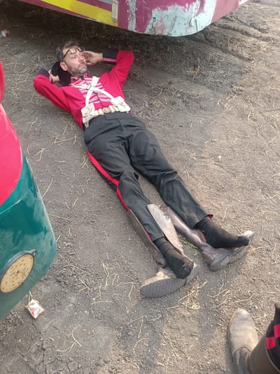 The scene is set outdoors in Kottalapalli, India, on dry, dark, textured ground consisting of small aggregate and dust, interspersed with light-colored dry grass or straw. The primary subject is a male individual, appearing middle-aged with a beard, mustache, and dark hair, lying supine on the ground. He wears spectacles pushed onto his forehead. He is dressed in a red, long-sleeved, military-style jacket with black cuffs, adorned with white cross-straps featuring multiple white pouches and a white belt with a gold-toned buckle. His lower body is clad in black trousers with a distinct red stripe running down each outer seam. His feet are covered in black socks. His hands are clasped behind his head, and his elbows rest on the ground, suggesting a resting posture. A dark, possibly black, hat or headpiece is held in his left hand, behind his head. Two worn, knee-high brown boots with visible soles are positioned on the ground near his feet, indicating they have been removed.
Portions of at least two vehicles are visible. A section of a vehicle with peeling paint, showing faded red and light green/blue colors, is present in the upper background, positioned over a rutted area of the ground. On the left foreground, part of another vehicle, painted red and a darker green, is visible, featuring a circular LED light fixture. A small, discarded red and white wrapper lies on the ground near this vehicle component. In the lower right corner, the lower leg and a dark-colored boot of another individual, with a visible red and black pattern near the top, are partially visible, indicating the presence of a second person out of frame. No direct interaction between individuals is depicted.