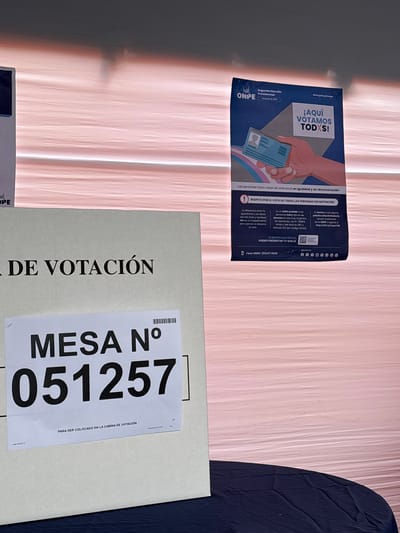 The image captures an indoor polling station environment in Santiago de Surco, Peru. A light-colored panel, likely part of a voting booth or ballot box, features a white label with "MESA N° 051257" printed prominently. Below the number, smaller text reads "PARA SER COLOCADO EN LA CABINA DE VOTACIÓN," with "LIMA OESTE 3" and "SANTIAGO DE SURCO" also visible. To the right, a blue and white poster from the Oficina Nacional de Procesos Electorales (ONPE) is affixed to a textured light pink wall or fabric. The poster, titled "Segunda Elección Presidencial 7 de Junio de 2025," displays the slogan "¡AQUÍ VOTAMOS TODXS!" (HERE WE ALL VOTE!) and an illustration of a hand holding an ID card over a background incorporating the trans pride flag colors. Text on the poster states, "Las personas trans votan en este local en igualdad y sin discriminación" (Trans persons vote in this polling station in equality and without discrimination) and "RESPETO POR EL VOTO DE TODAS LAS PERSONAS SIN DISTINCIÓN." Additional text provides legal information regarding the right to vote for trans individuals, prohibits discrimination, and lists a contact phone number (Fono ONPE: (01) 417 0830) and a website (www.gob.pe/onpe) for reporting grievances. A dark blue tablecloth covers a table in the foreground, indicating a surface below the voting equipment.
