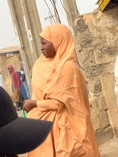 A woman with a dark complexion is depicted in a close-up, standing on a street in Muchiya, Nigeria. She wears a light orange-tan head covering (hijab) and a matching full-body garment. Her facial expression shows a slight frown or downturned mouth, and her gaze is directed towards the left. Her right hand is visible, holding a small green object or bag strap. In the immediate foreground, the brim of a dark baseball cap partially obscures the lower left portion of the frame.
The background reveals a bustling urban environment. To the left, several utility poles or concrete pillars are visible, adorned with various posters or notices. Behind these poles, a small, light-colored structure or stall is present, with the words "HAFIDU OIL KERO" legible on its side, indicating a commercial establishment. A young individual in a red hooded top and a long brown garment stands near this structure, looking towards the camera. Other indistinct figures are partially visible further back. To the right of the main subject, a section of an old, weathered wall made of light-colored, possibly mud-rendered stone, shows significant cracks and texture. Overhead, multiple utility wires and cables traverse the scene against a hazy, light sky. The ground appears to be dry earth or dust.
